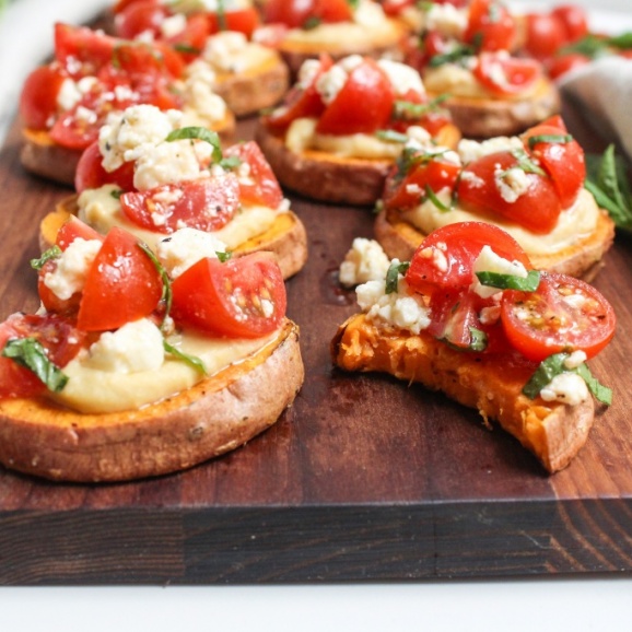 slices of sweet potato topped with tomatoes and hummus on a cutting board